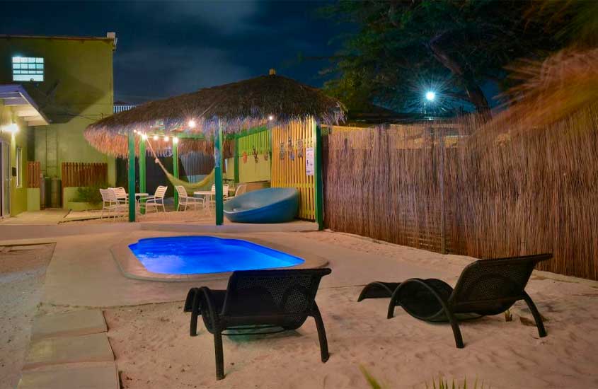 Durante a noite, área de lazer de hotel em aruba com espreguiçadeiras, banco, mesas, cadeiras, piscina e parte de areia