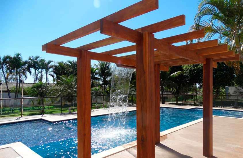 Em um dia de sol, área de lazer de hotel em bonito com piscina, cascata, estrutura de madeira árvores e parte gramada ao redor