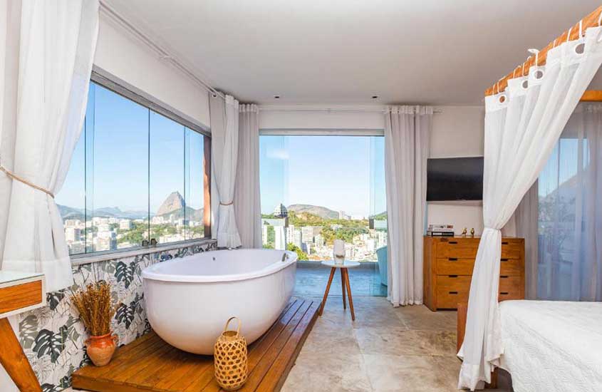 Quarto de um dos hotéis que aceitam pet no Rio de Janeiro com cama de casal, cômoda, banheira, TV, janelas grandes acortinadas, vasos decorativos e vista da cidade
