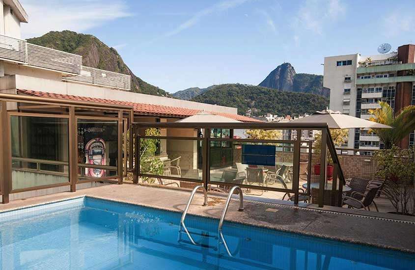 Em dia de sol, área de lazer de um dos hotéis que aceitam pet no Rio de Janeiro com piscina, mesas, cadeiras, freezer, plantas decorativas e vista do pão de açúcar
