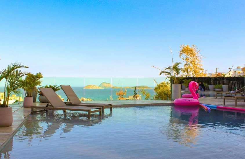Em final de tarde, área de lazer de um dos hotéis que aceitam pet no Rio de Janeiro com piscina, boia, espreguiçadeiras, vista para o mar e plantas decorativas