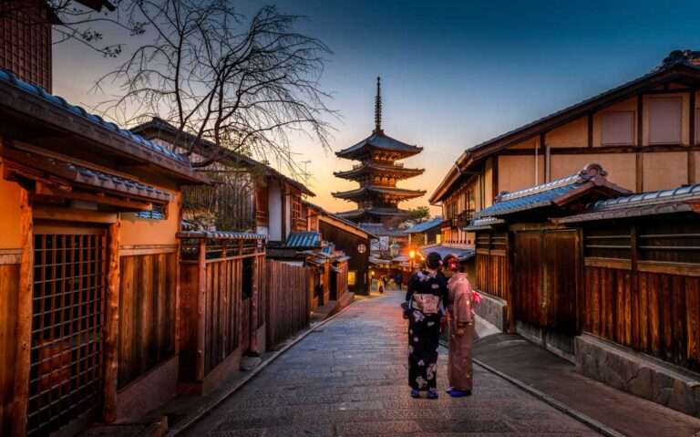 Em um final de tarde em kyoto japão, duas mulheres caminham em rua iluminada com luzes amarelas, árvore seca e vista para um templo