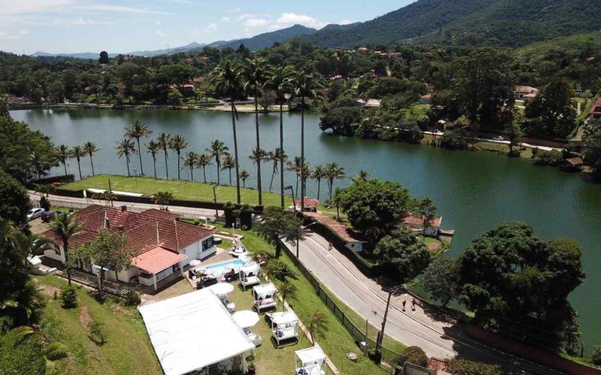 Em um dia de sol, vista aérea de prédio, piscina e tendas em área de hotel fazenda em Miguel Pereira com vista para lago rodeado por árvores