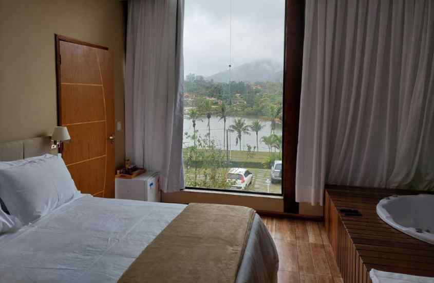 Em um dia nublado, quarto de hotel com cama de casal, frigobar, janela grade janela acortinada, deck de madeira e banheira de hidromassagem