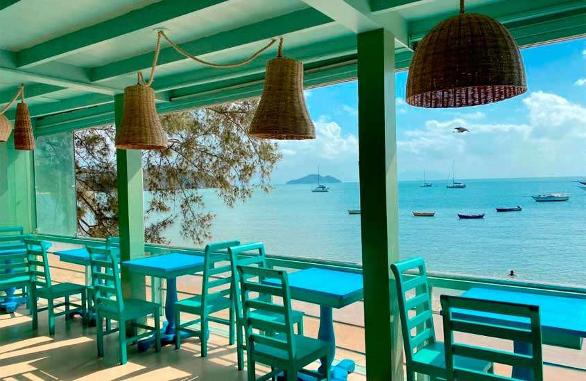 Em um dia de sol, área de café da manhã de um dos hostels em búzios com mesas, cadeiras, luminárias de palha e mar com barcos na frente