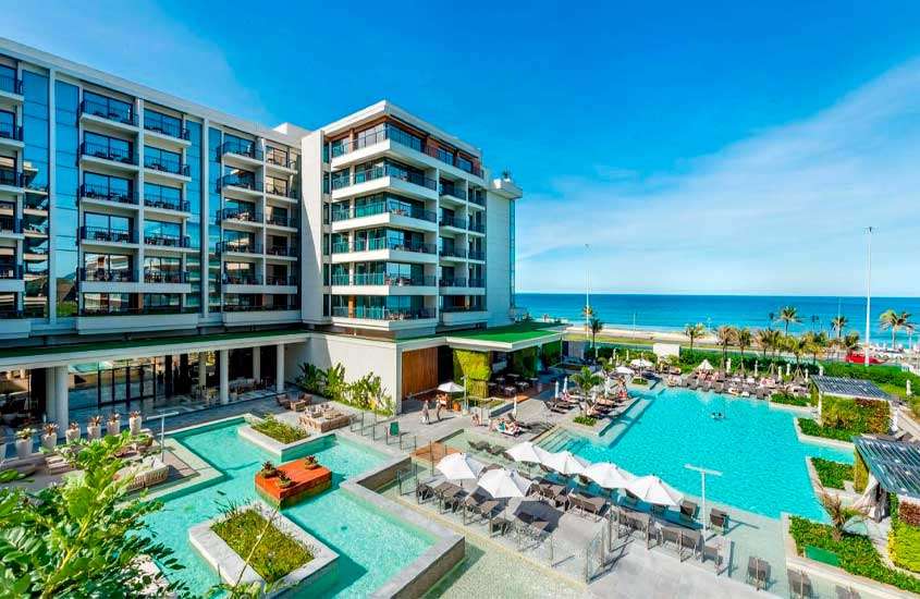 Em um dia ensolarado, vista aérea de um dos hotéis na Barra da Tijuca em frente à praia com piscinas, plantas decorativas, espreguiçadeiras, guarda-sóis, pessoas e árvores