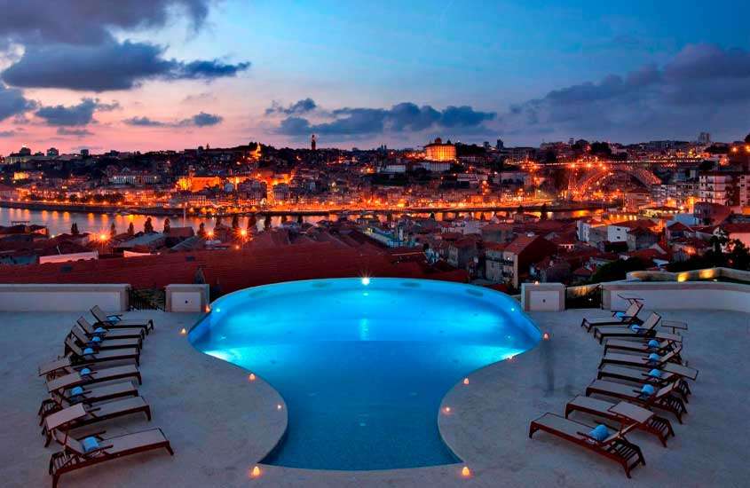 Durante o anoitecer, área de lazer de um dos melhores hoteis vinicolas portugal com piscina, espreguiçadeiras de madeira, toalhas, cidade iluminada por luzes amarelas e rio na frente