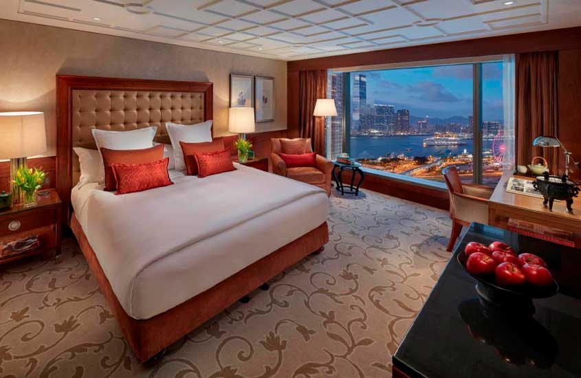Quarto de um dos melhores hotéis em Hong Kong com cama de casal, carpete, poltrona, mesa, área de trabalho, luminárias, maçãs, quadros decorativos e janelas grandes acortinadas
