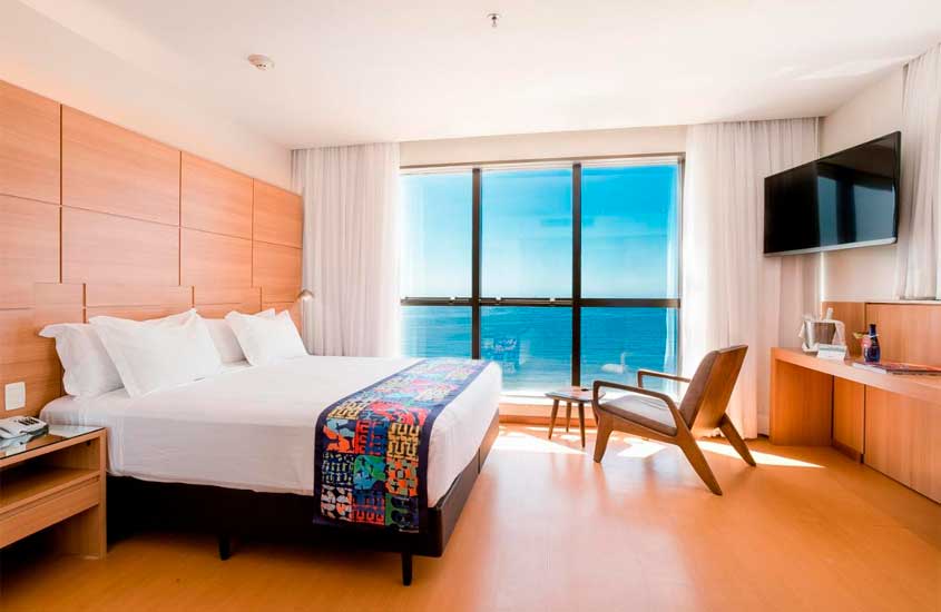 Em um dia de sol, quarto de um dos hotéis em copacabana com vista para o mar com cama de casal, poltrona, criado, mesa, TV e janela grande acortinada