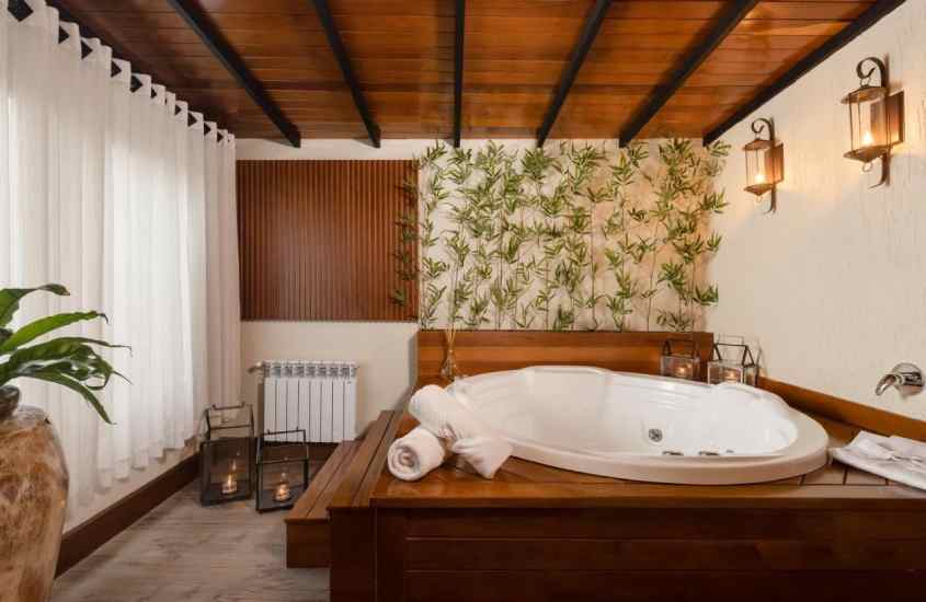 Banheiro de um hotel em gramado com banheira, aquecedor, toalhas e janela grande acortinada