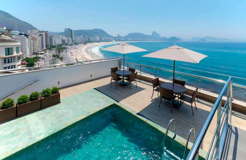 Durante um dia de sol, área de lazer de um hotel em copacabana frente mar com mesas, cadeiras, guarda-sóis, flores decorativas, piscina, mar e cidade ao lado