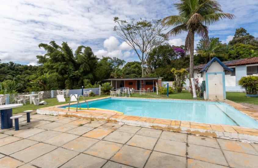 Em um dia de sol com nuvens, área de lazer de hotel fazenda em Miguel Pereira com piscina, mesas, cadeiras, bancos e área verde ao redor
