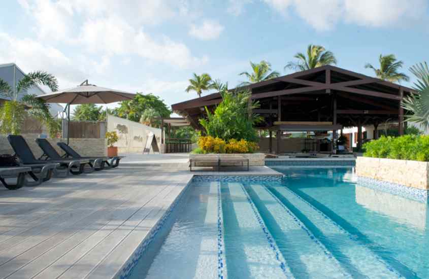 Em um dia de sol, área de lazer de um resort em curaçao com piscina, espreguiçadeiras, ombrelone e plantas decorativas