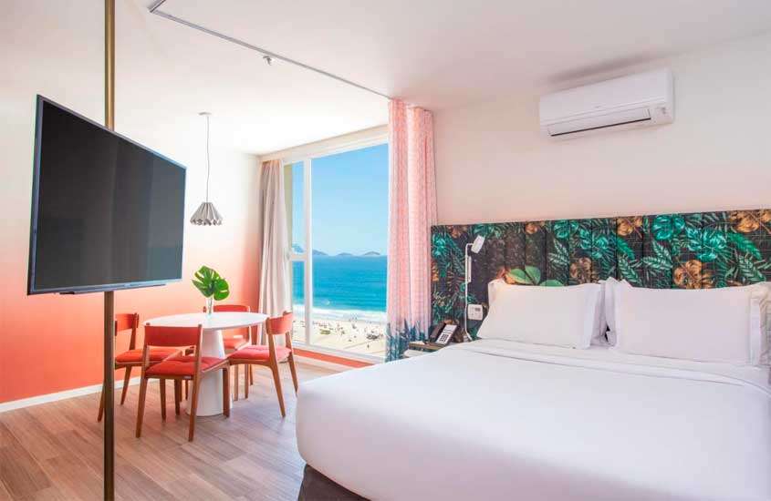 Quarto de um hotel em copacabana de frente para o mar com cama de casal, TV, mesa, cadeiras, ar-condicionado, luminária, flor decorativa e janela grande acortinada