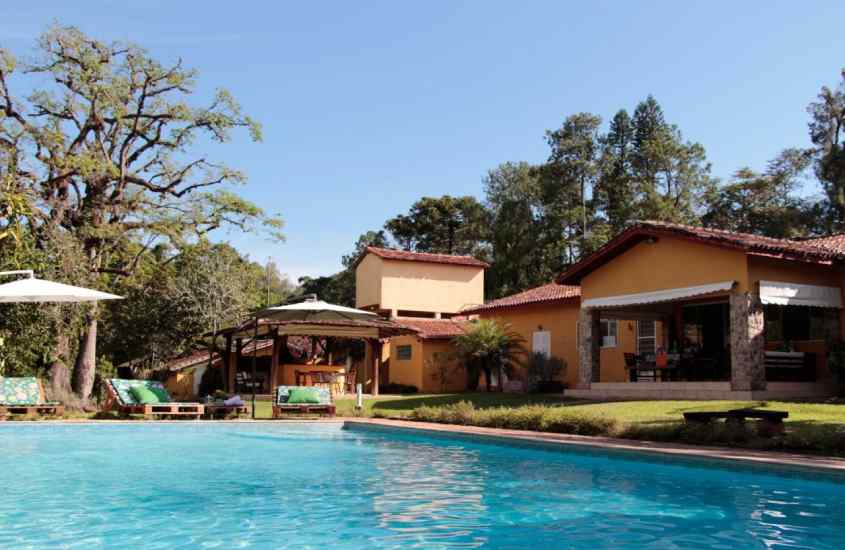 Em um dia de sol, área de lazer de um hotel fazenda em Ribeirão Preto com piscina, espreguiçadeiras, parte gramada, ombrelone e árvores grandes ao redor