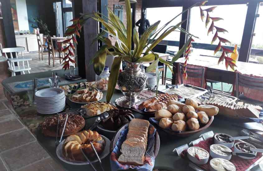 Mesa de café da manhã com bolos, pães, geleias, manteiga, frios e flor decorativa