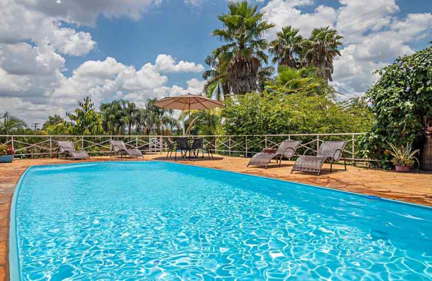 Em um dia de sol com nuvens, área de lazer de um hotel fazenda em Ribeirão Preto com piscina, espreguiçadeiras, mesa, cadeiras e vegetação ao redor