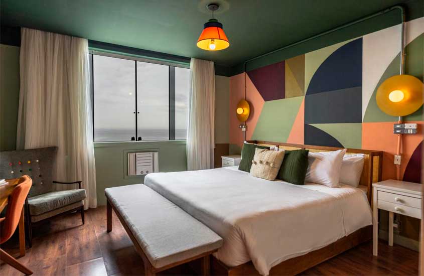 Quarto de um dos hotéis em copacabana beira mar com cama de casal, poltrona, cadeira, mesas, luminárias, ar-condicionado, parede colorida e criados