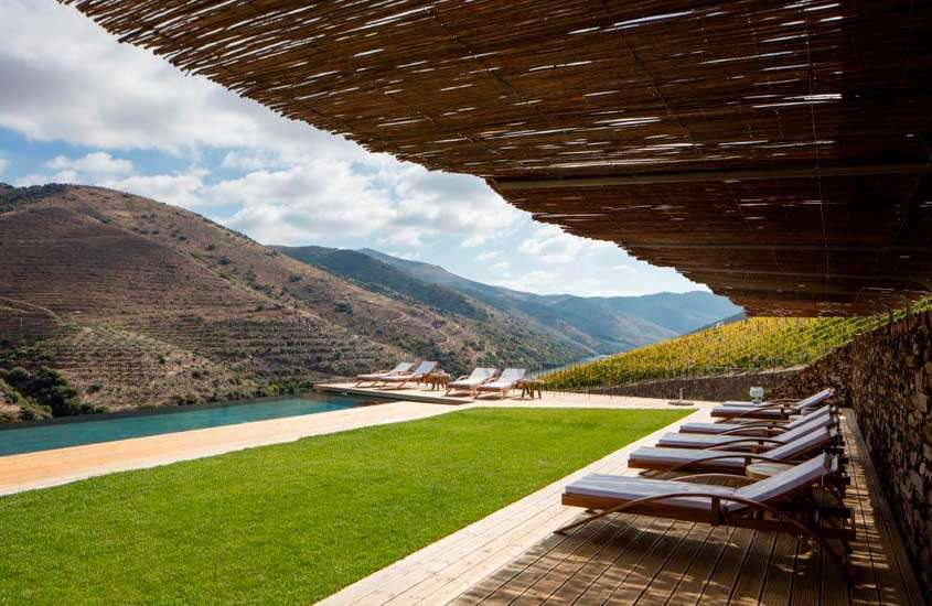 Em um dia de sol, área de lazer coberta de um dos hoteis vinícolas em portugal com espreguiçadeiras de madeira, parte gramada, piscina e plantações de uva ao redor