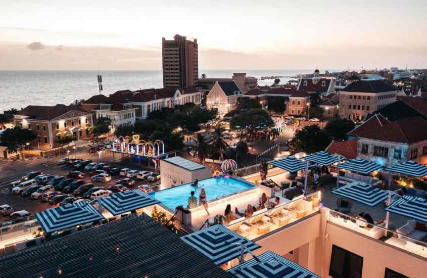 Durante o anoitecer, vista aérea da cidade com piscina na cobertura do hotel, árvores e prédios ao redor, e carros estacionados