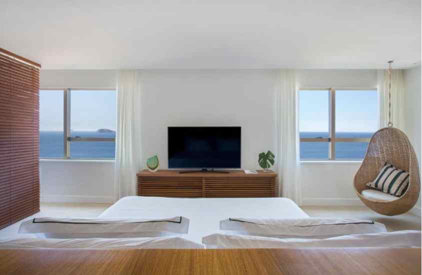 Quarto de um dos hoteis no rio de janeiro com vista para o mar, cama de casal, cadeira suspensa, TV e janelas grandres