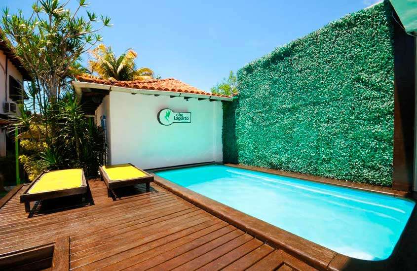 Em um dia de sol, área de lazer de um hostel em búzios com piscina, parede de grama, espreguiçadeiras e deck de madeira e plantas decorativas
