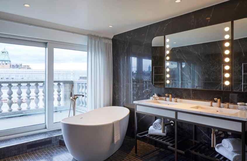Banheiro de hotel com espelho camarim, banheira, toalhas e janela acortinada