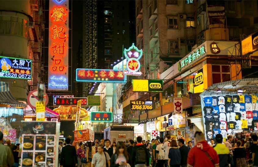 Durante a noite, bairro onde ficar em Hong Kong com letreiros iluminados, pessoas e prédios.