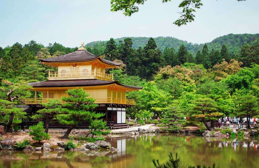 Em um dia de sol, templo em kyoto com andares dourados, lago ao redor, vegetação e visitantes