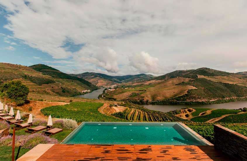 Em um dia de sol, área de lazer de um dos melhores hoteis vinicolas portugal com espreguiçadeiras de madeira, guada-sóis, plantações e rio atrás