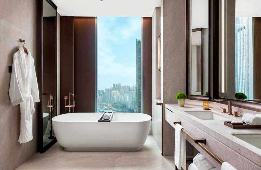 Banheiro de um hotel em Hong Kong com banheira, toalhas, pia, espelhos, janela grande acortinada e planta decorativa