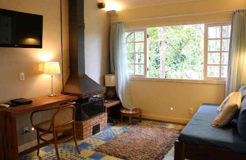 Sala de uma acomodação de hotel fazenda em friburgo com sofá, tapete, lareira, TV, área de trabalho e janela grande acortinada