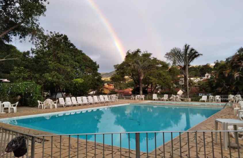 Em um dia nublado, área de lazer de um hotel com piscina, mesas, cadeiras, espreguiçadeiras, árvores e arco-íris atrás