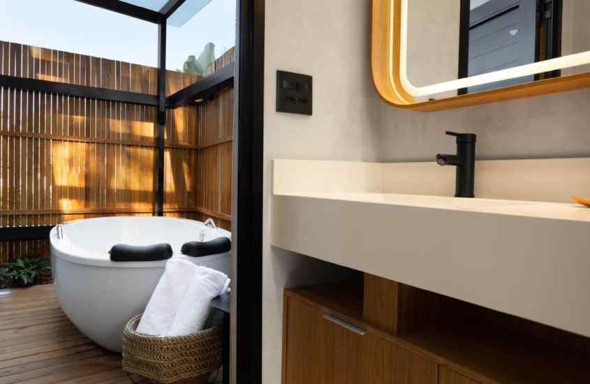 Banheiro com banheira, pia grande, toalhas, deck de madeira e espelho iluminado