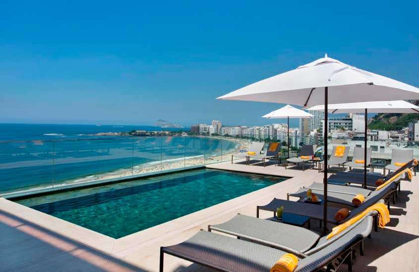 Em um dia ensolarado, área de lazer de um hotel em copacabana perto da praia com piscina de borda infinita, espreguiçadeiras, toalhas, guarda-sóis e litoral na frente