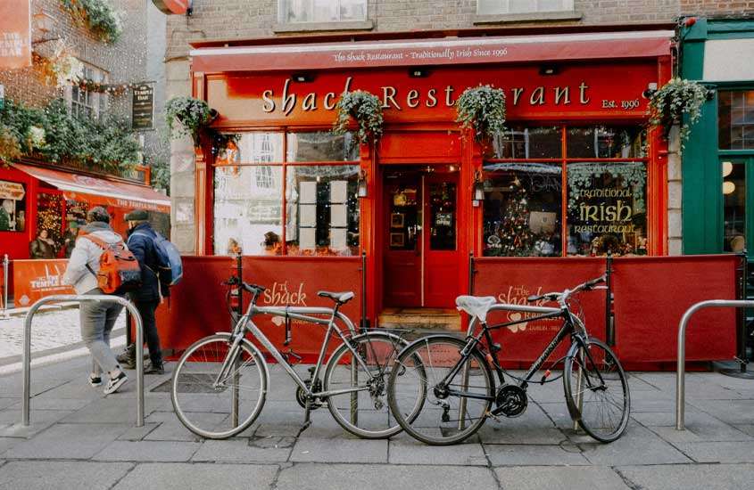 Durante o dia, fachada de um restaurante vermelho com plantas decorativas, bicicletas e pessoas na frente
