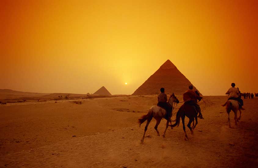Durante o pôr do sol, pessoas cavalgando em direção as pirâmides
