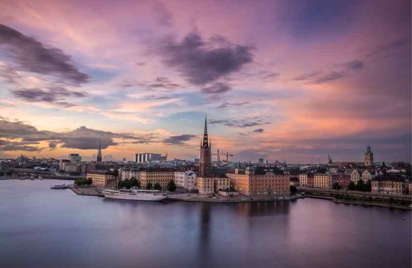 Durante o entardecer, vista aérea da cidade de Estocolmo com mar na frente refletindo o céu e prédios grandes ao fundo