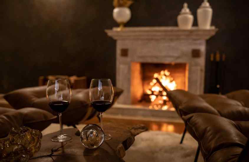 Área de descanso com poltronas, lareira a lenha, tapete felpudo e taças de vinho tinto