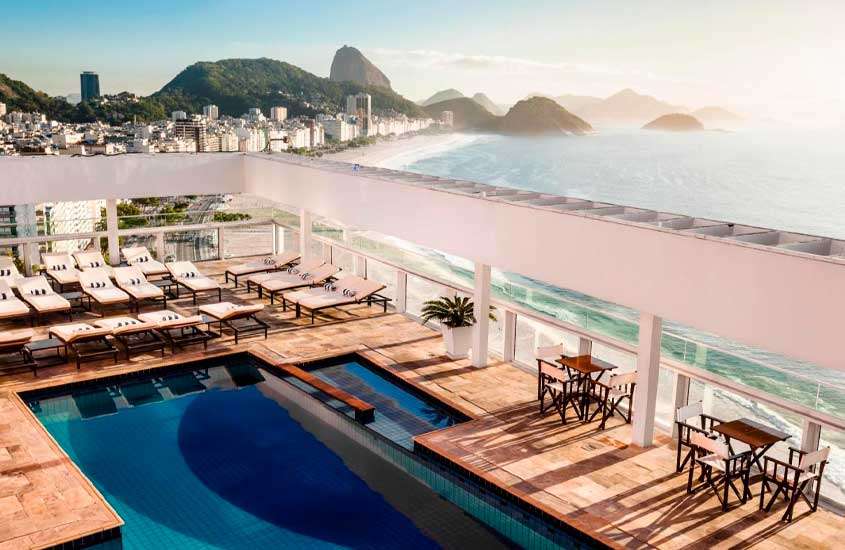 Em um dia de sol, área de lazer de um dos hotéis em copacabana beira-mar com espreguiçadeiras, mesas, cadeiras, piscina, flores decorativas, mar e cidade ao redor