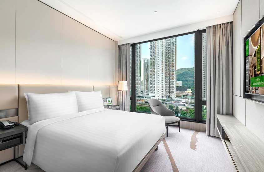 Quarto de hotel onde ficar em Hong Kong com cama de casal, poltrona, TV, luminária, criado e janela grande acortinada