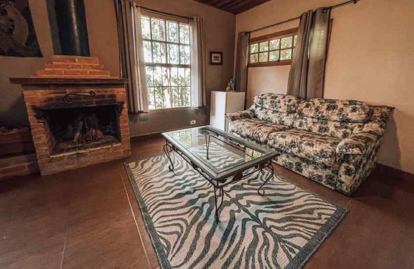 Sala de estar de um dos chalés românticos em são roque com sofá, mesa, lareira de tijolos, tapete, janelas grandes acortinadas e quadro decorativo