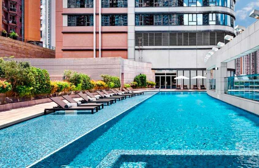 Em um dia de sol, área de lazer de um hotel onde ficar em Hong Kong com piscina, espreguiçadeiras, toalhas, plantas decorativas, guarda-sóis e hotel ao redor