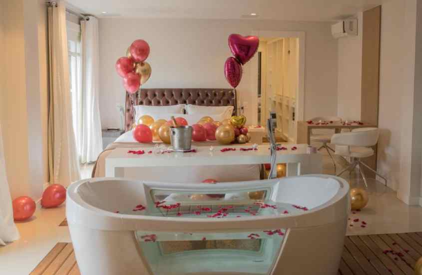 Quarto de hotel em gramado com banheira e cama de casal, ambas decoradas com com pétalas de rosa e balões, criando um clima romântico
