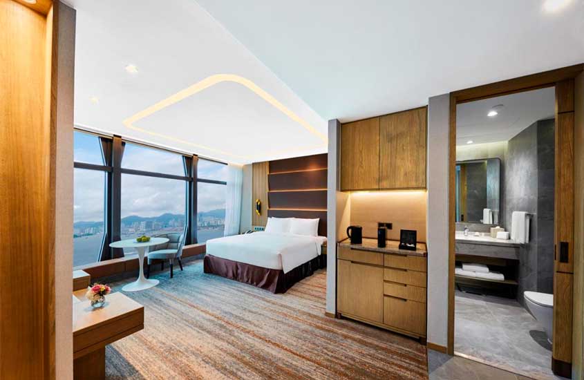 Quarto de um dos melhores hotéis em Hong Kong com banheiro, área do café, cama de casal, painel de madeira, poltrona, mesa e janela grande acortinada