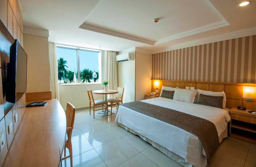 Quarto de um hotel em copacabana com vista para o mar com cama de casal, cadeiras, mesa, TV, ar-condicionado, luminárias e janela acortinada
