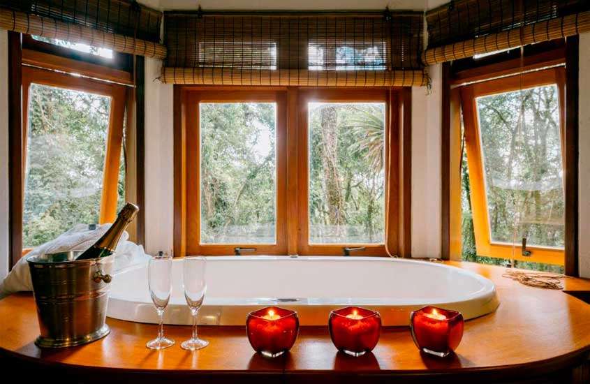 Banheira de um dos chalés em monte verde com hidro, velas, taças e balde de champagne e janelas grandes de madeira