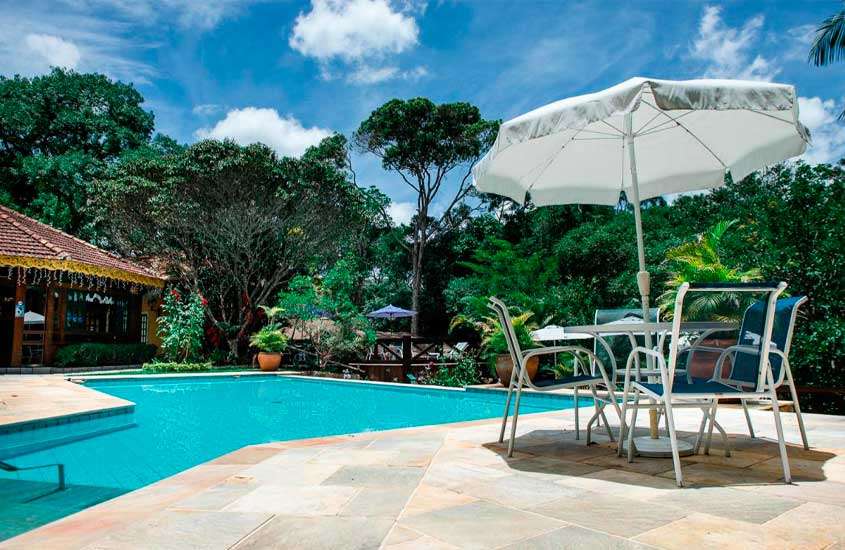 Em um dia de sol, área de lazer de um hotel em teresópolis com piscina, mesa, cadeiras, guarda-sol e árvores ao redor