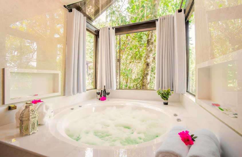 Banheira de um dos chalés em monte verde com hidro, janelas grandes acortinada e plantas decorativas