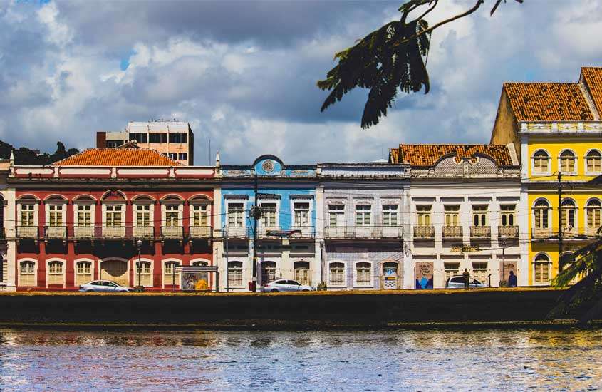 Durante um dia de sol com nuvens, orla de Recife com casas coloridas atrás e árvore ao lado